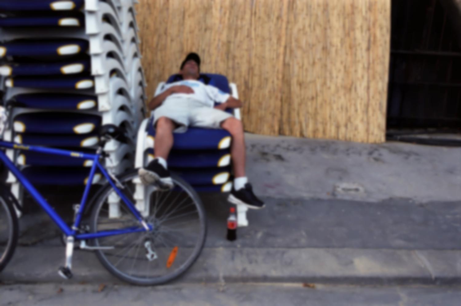 Homme allongé sur une pile de chaises longues, le pied appuyé sur la roue d'un vélo. Paris, août 2003.