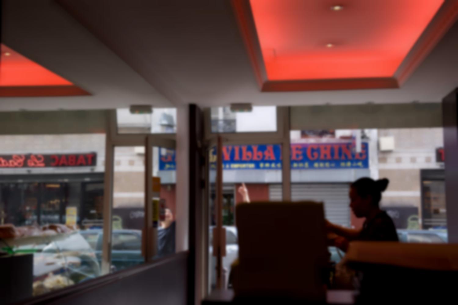 Cantine chinoise rue de Joinville, la serveuse en contrejour, un miroir qui reflète la vitrine. Dans la rue, un homme montre quelque chose en l'air, et l'enseigne d'un autre restaurant, la Villa de Chine. Paris, août 2011.