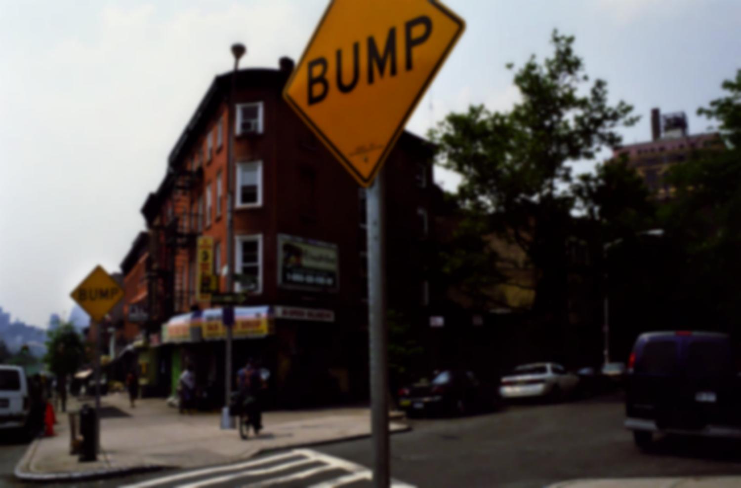 A l'entrée d'un passage piétons, près d'immeubles de briques, un panneau carré jaune : BUMP. New York, juin 2003.