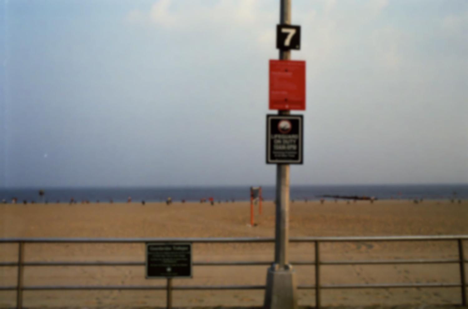 Brighton Beach : devant la plage et la mer, accrochés à une barrière et un poteau, un règlement, et, en anglais et en espagnol, les horaires de présence des sauveteurs - interdiction de se baigner en-dehors de ces heures. New York, juin 2003.