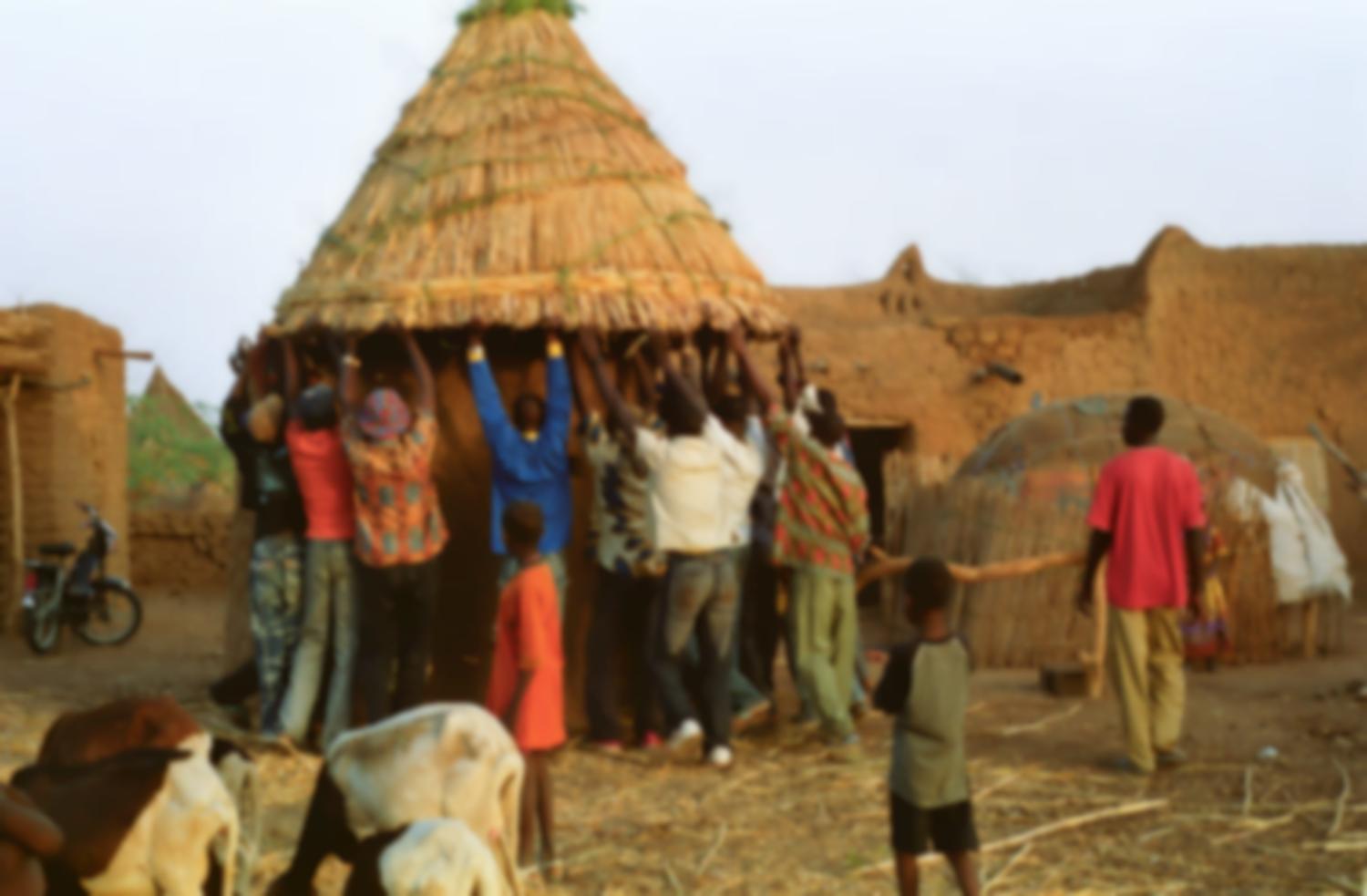 Des hommes soulèvent un toit conique fait de mil pour le poser sur un petit édifice circulaire : une cuisine. Bosseye Dogabe, Burkina Faso, mai 2008.