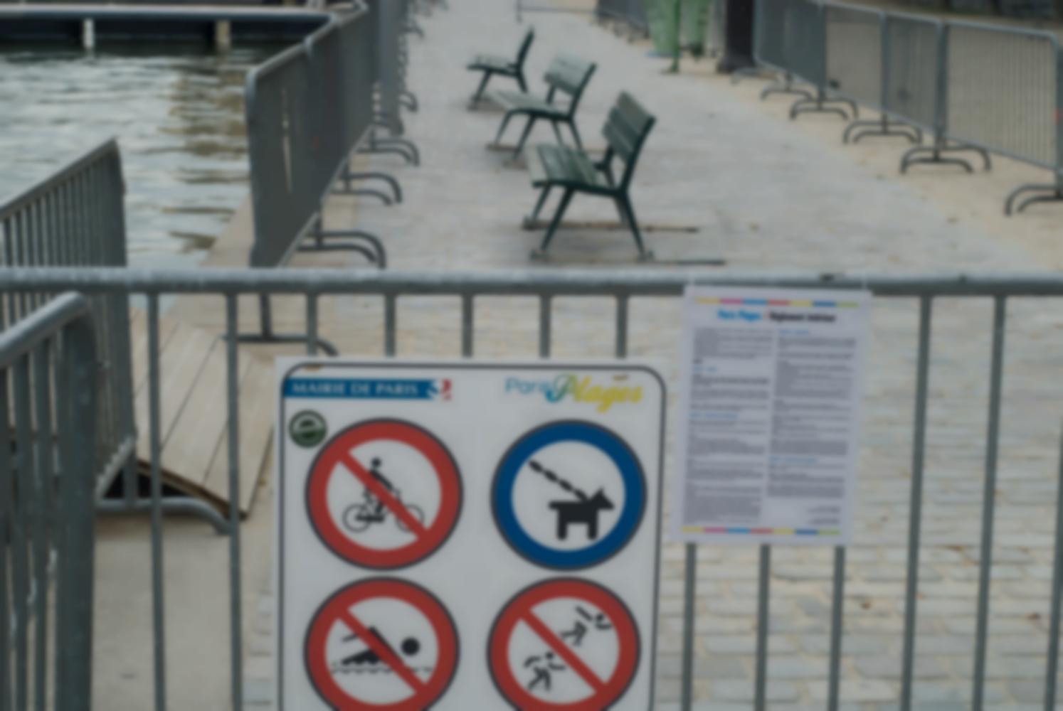 Des barrières métalliques encerclent trois bancs et les séparent de l'eau. Sur l'une d'elles est accroché le règlement de Paris Plage, dont quatre interdictions sont soulignées par des pictogrammes. Paris, août 2010.