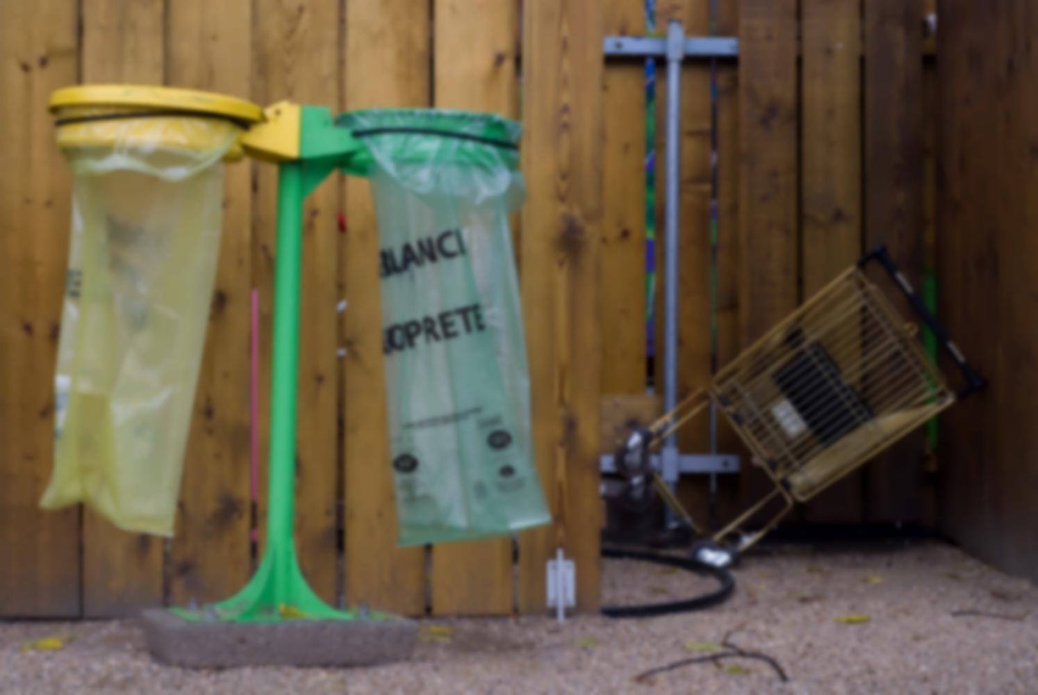 Devant une palissade en lattes de bois, une double poubelle, jaune et verte : deux sacs plastique et leur support, pour le recyclage. Derrière, dans un recoin, un caddie renversé. Paris, août 2010.