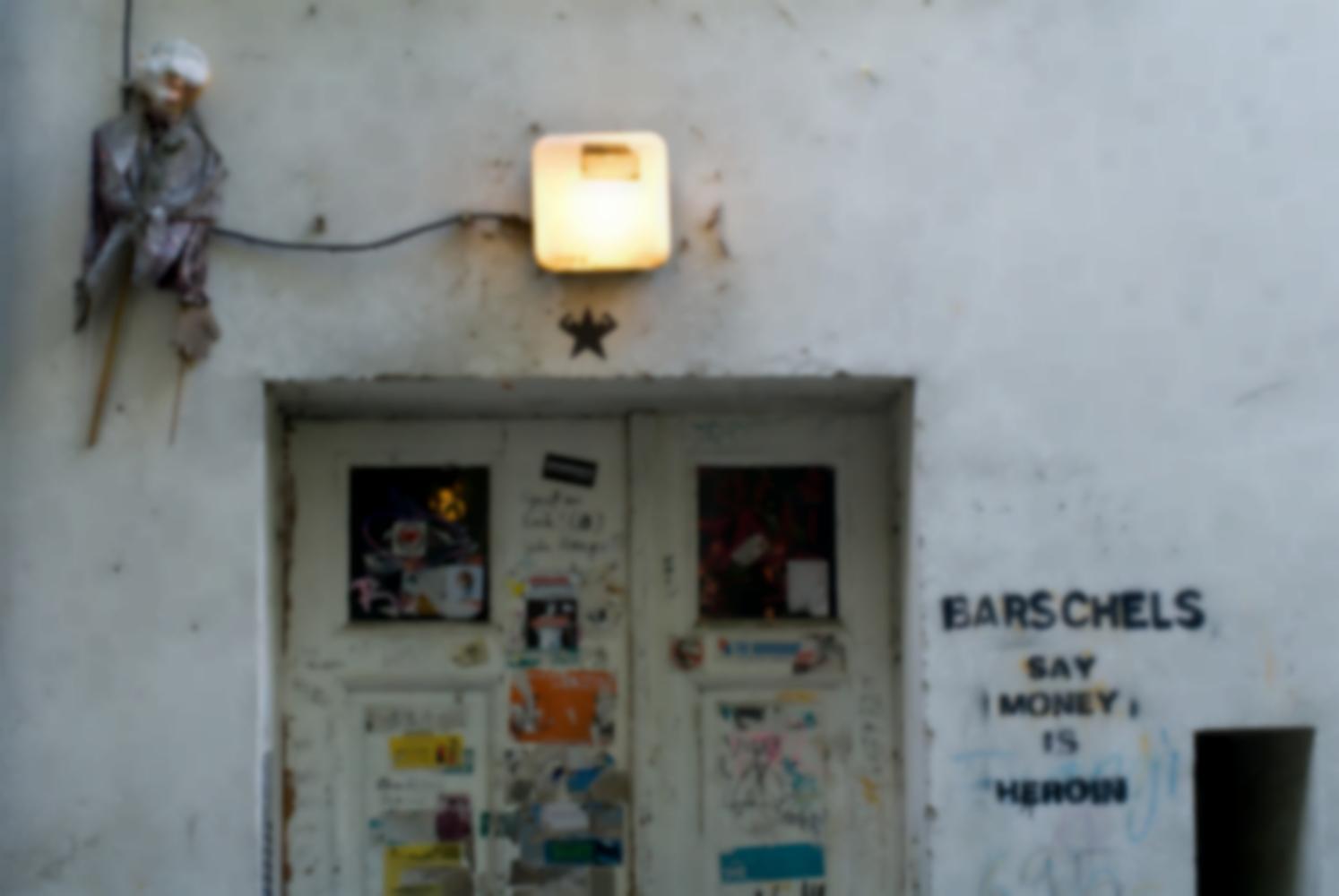 A côté d'une double porte couverte d'affiches et d'autocollants déchirés, surmontée d'une lumière et d'une sorte de grande marionnette, un graffiti au pochoir cite Barschels. Berlin, septembre 2009.