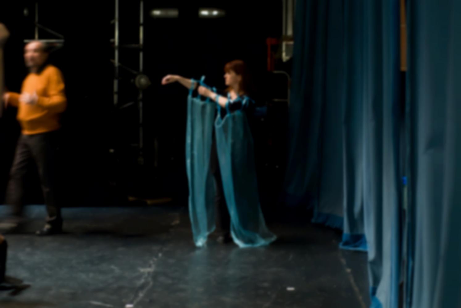 La scène avant la représentation, les rideaux sont fermés, une comédienne arrange deux voiles bleus attachés à ses bras par des rubans, près d'un comédien en pull-over orange. Nanterre, octobre 2009.