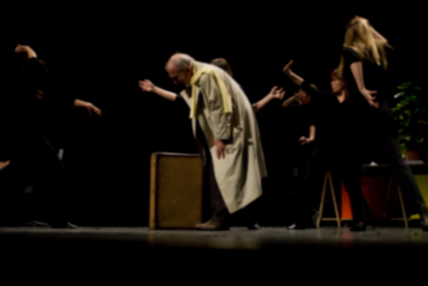 Deux bras d'une choreute dépassent derrière le père en imperméable, penché près de sa valise, autour duquel le chœur évolue d'une façon menaçante. Nanterre, octobre 2009.