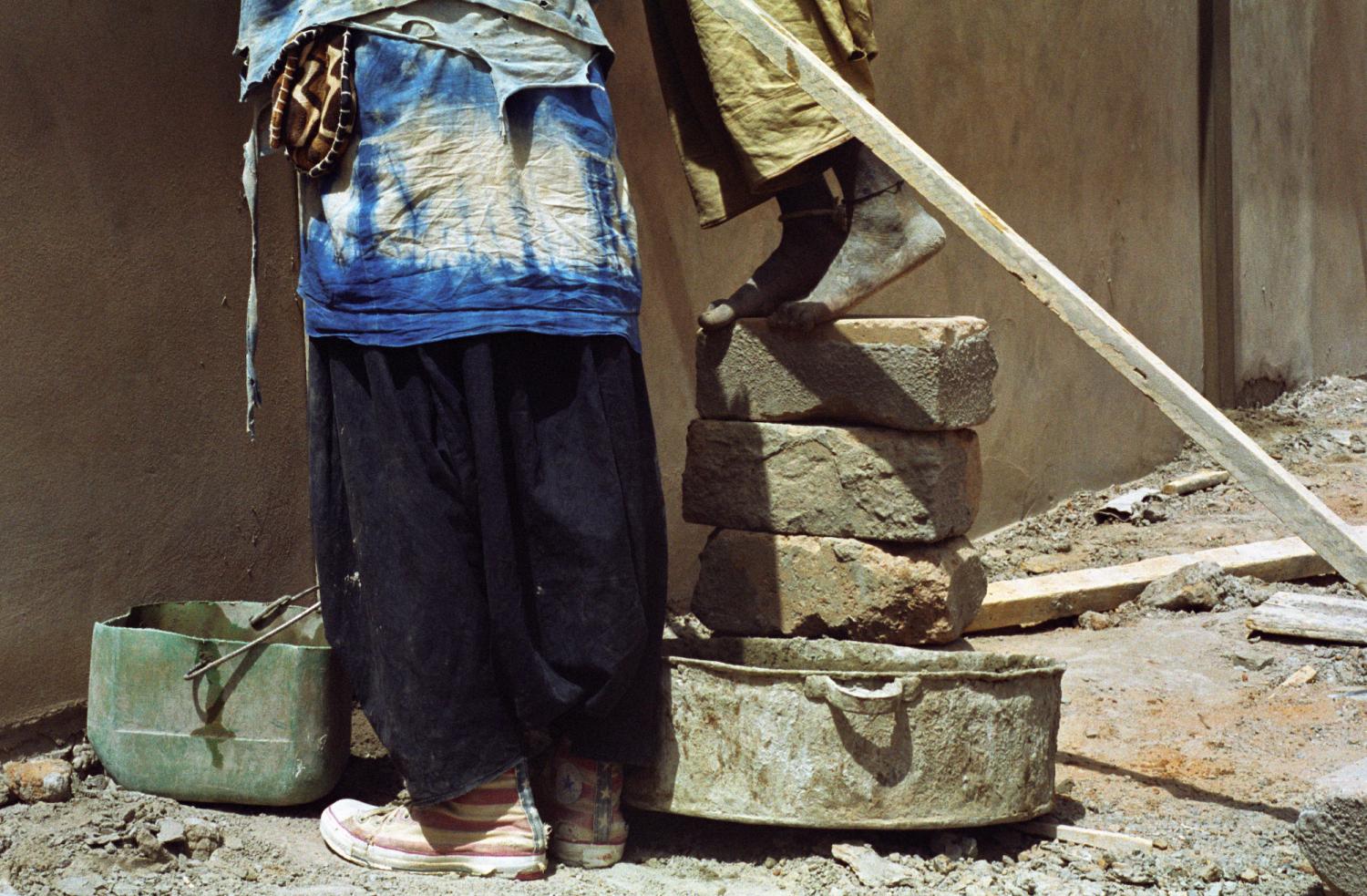 Jambes et pieds de Baye Fall au travail : l'un a des des baskets américaines, l'autre, monté sur une pile de parpaings, est sur la pointe de ses pieds nus. Touba, Sénégal, février 2008.