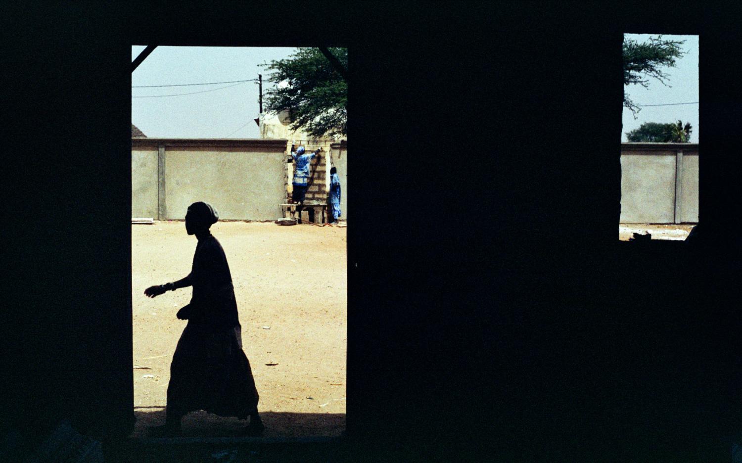 Un Baye Fall passe en contrejour devant la porte d'une pièce en construction. Plus loin, d'autres Baye Fall travaillent sur un mur. Touba, Sénégal, février 2008.