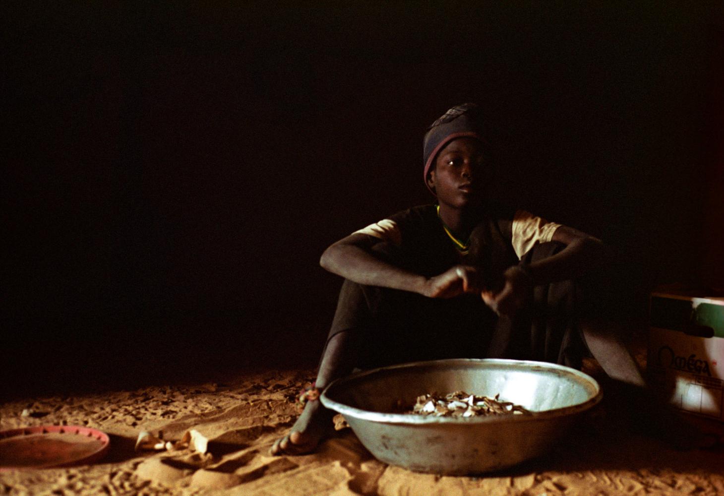 Assis sur le sol de sable, une grande bassine métallique devant lui, Aliou découpe du carton en petits morceaux. Touba, Sénégal, février 2008.