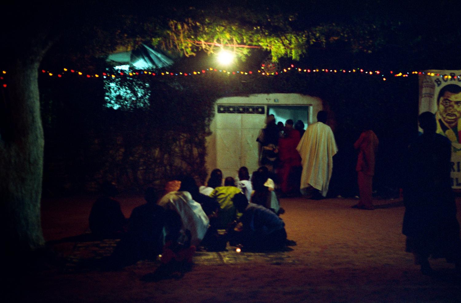 Devant la maison du marabout, sous un arbre, éclairé par une ampoule et une guirlande lumineuse, un groupe de pèlerins assis en cercle, tandis que d'autres invités entrent dans la concession. Touba, Sénégal, février 2008.