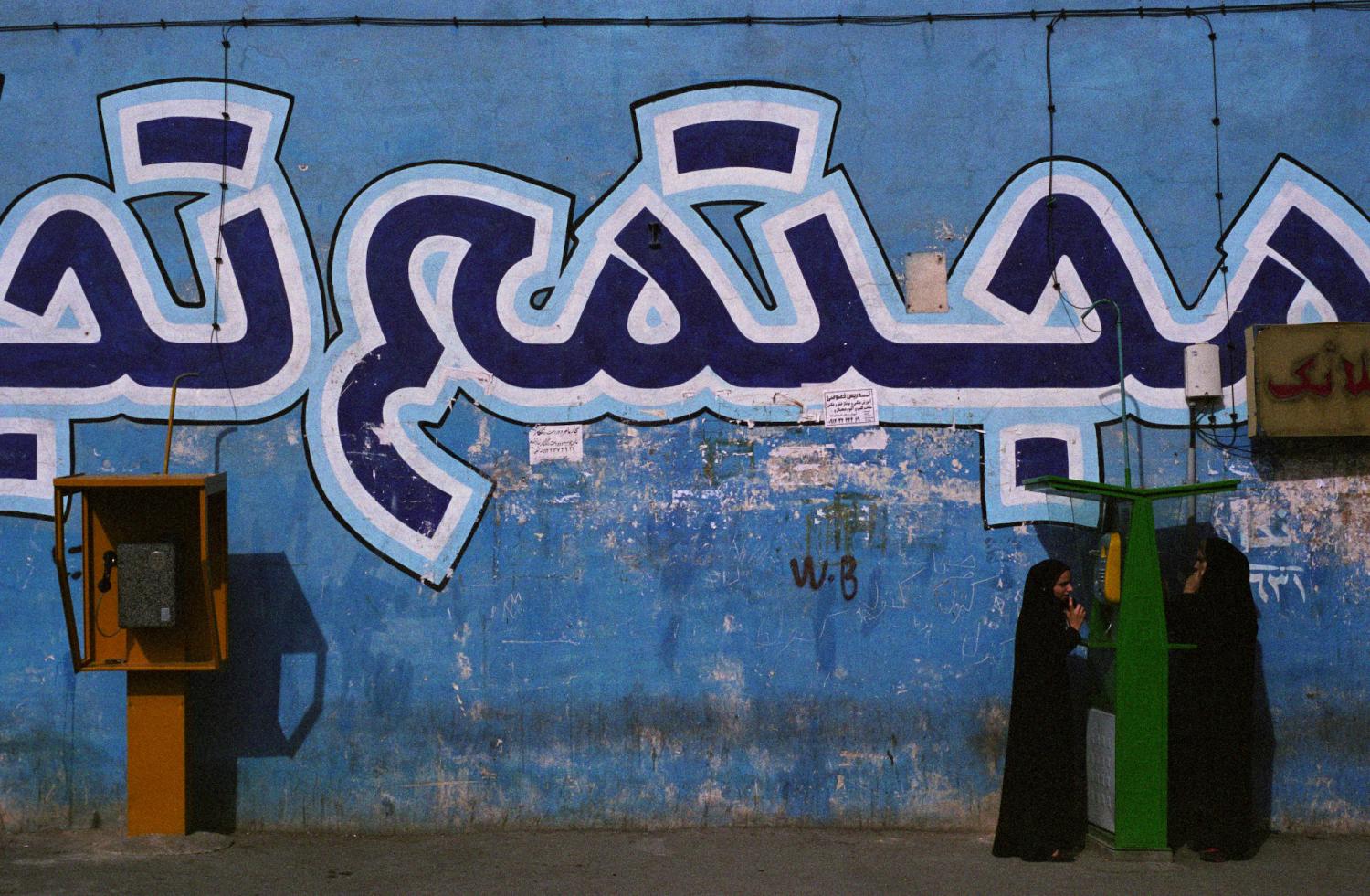Deux femmes en tchadors téléphonent à une cabine publique verte, près d'une autre jaune, devant un mur bleu décoré de grandes inscriptions en persan. Shahr-e-Rey, Iran, mars 2007.
