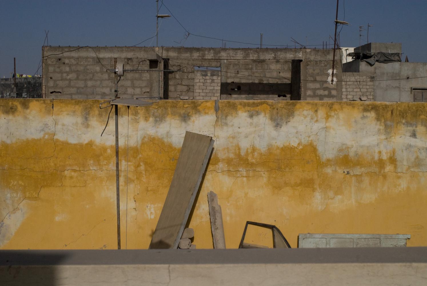 Derrière le muret du toit, un mur jaune auquel sont appuyés une planche, une portière de voiture, une porte de bois. Au-dessus dépasse un immeuble en construction. Dakar, Sénégal, mars 2010.