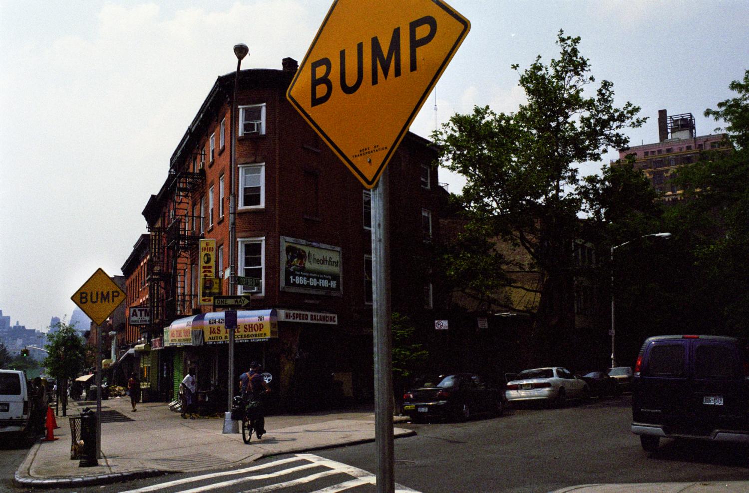 A l'entrée d'un passage piétons, près d'immeubles de briques, un panneau carré jaune : BUMP. New York, juin 2003.