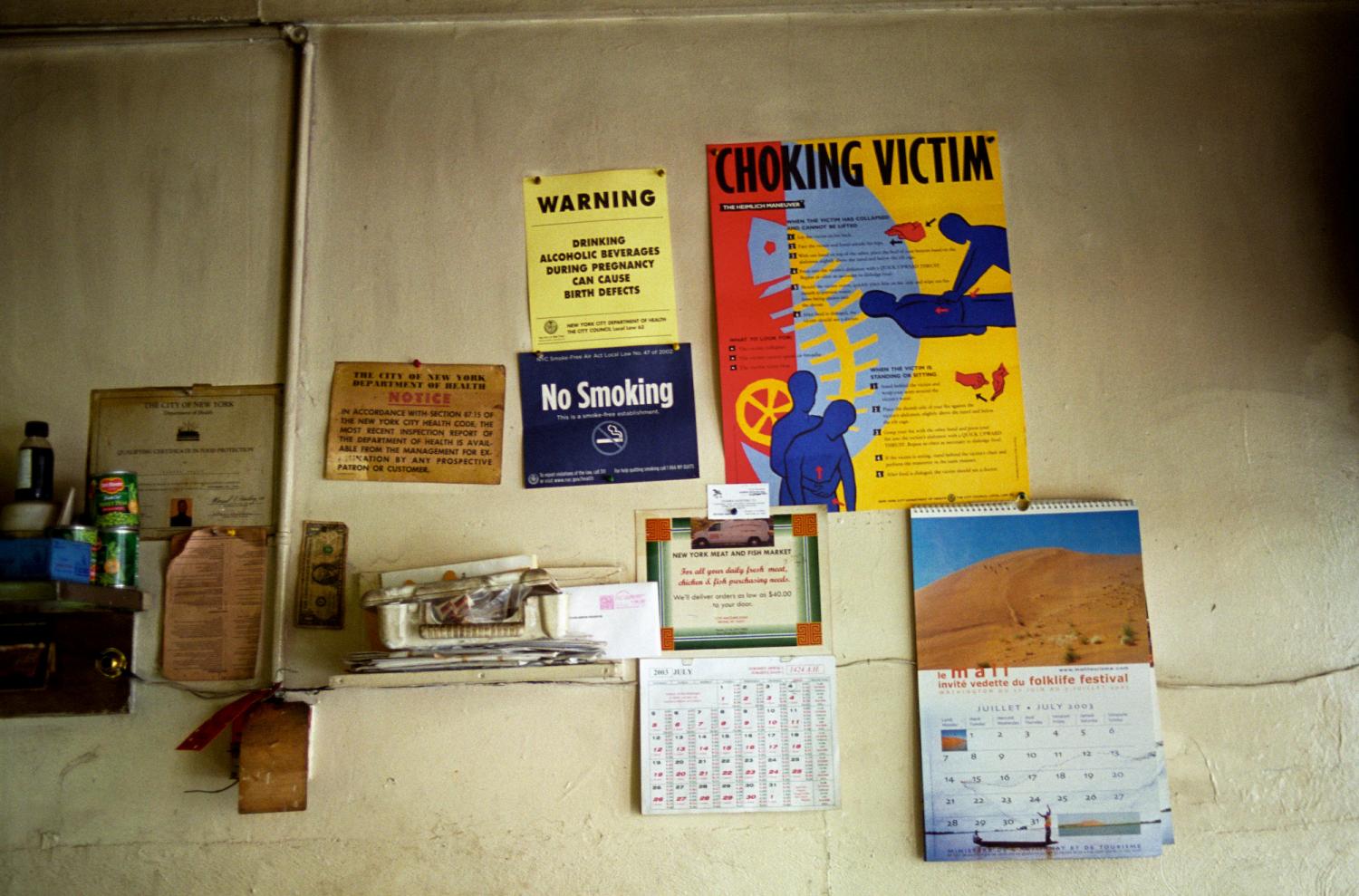 Sur le mur d'un restaurant, parmi divers objets, des avertissements sur les dangers de l'alcool pendant la grossesse, une interdiction de fumer, des instructions de réanimation et des informations sur l'inspection sanitaire. New York, juillet 2003.