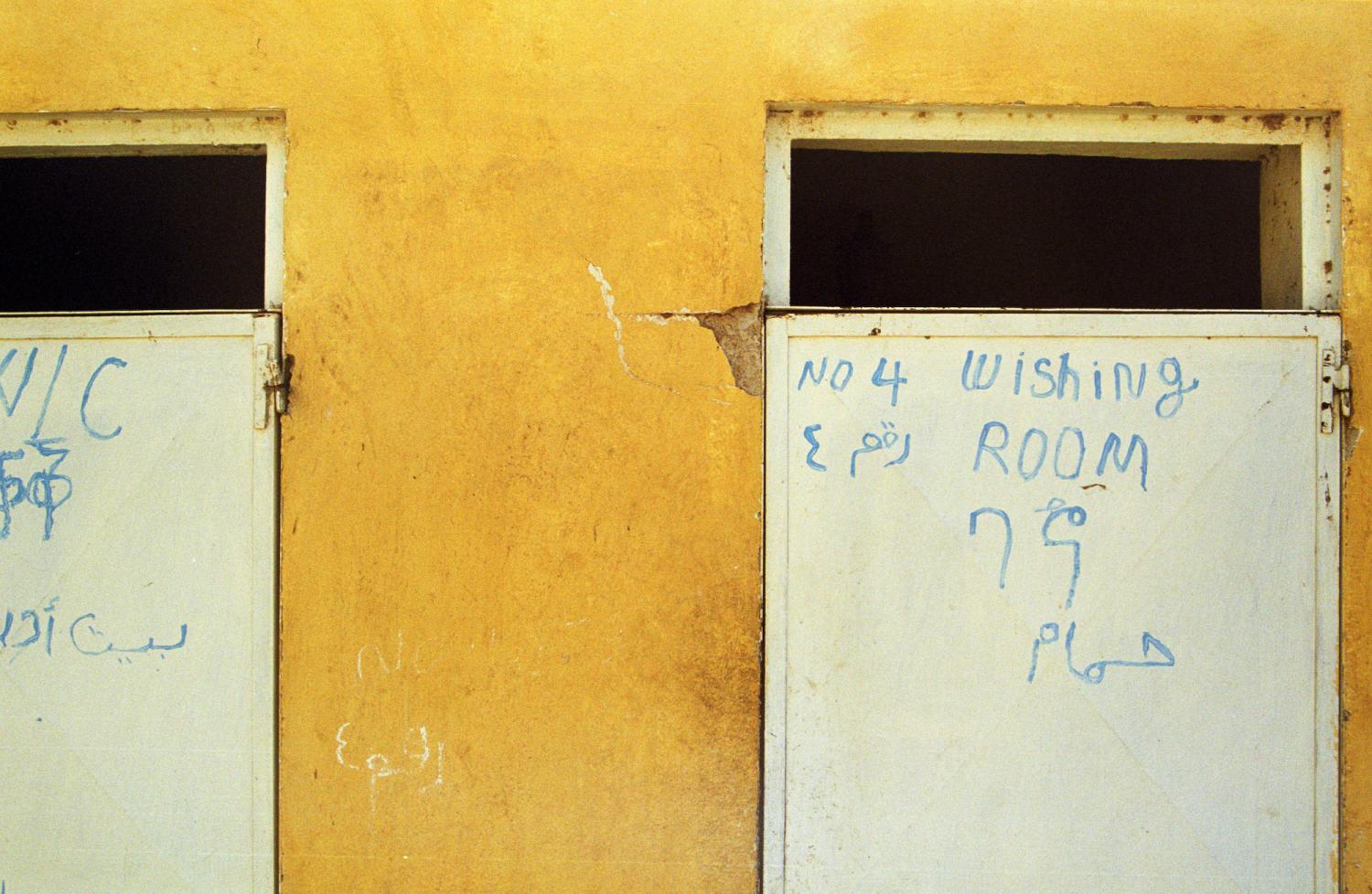 Dans un mur jaune, deux portes blanches avec inscriptions en plusieurs langues et alphabets - dont WC et Wishing room. Teseney, Erythrée, février 2005.
