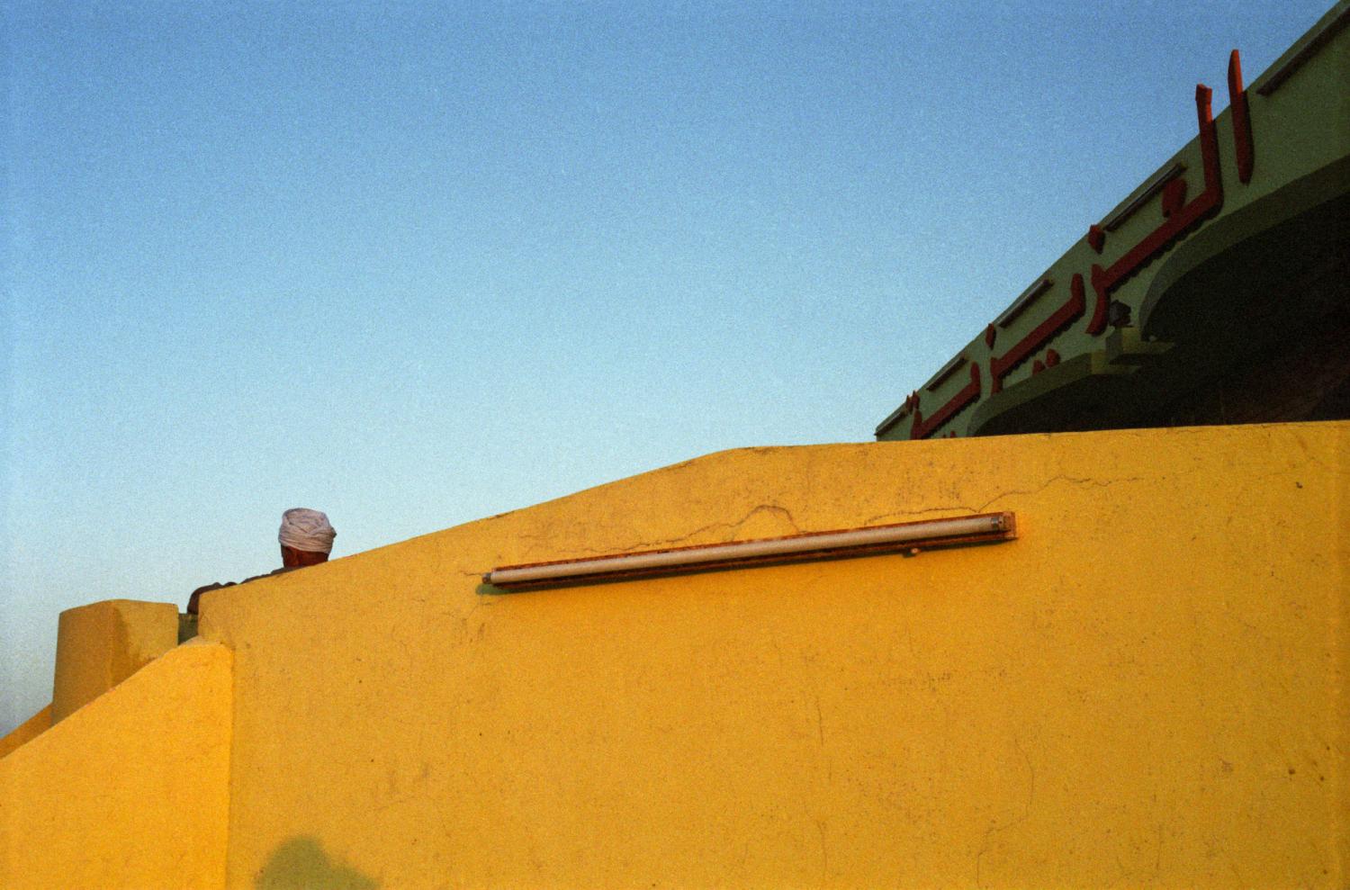 Une tête enturbannée dépasse du mur jaune de la terrasse d'un restaurant routier. Egypte, avril 2004.