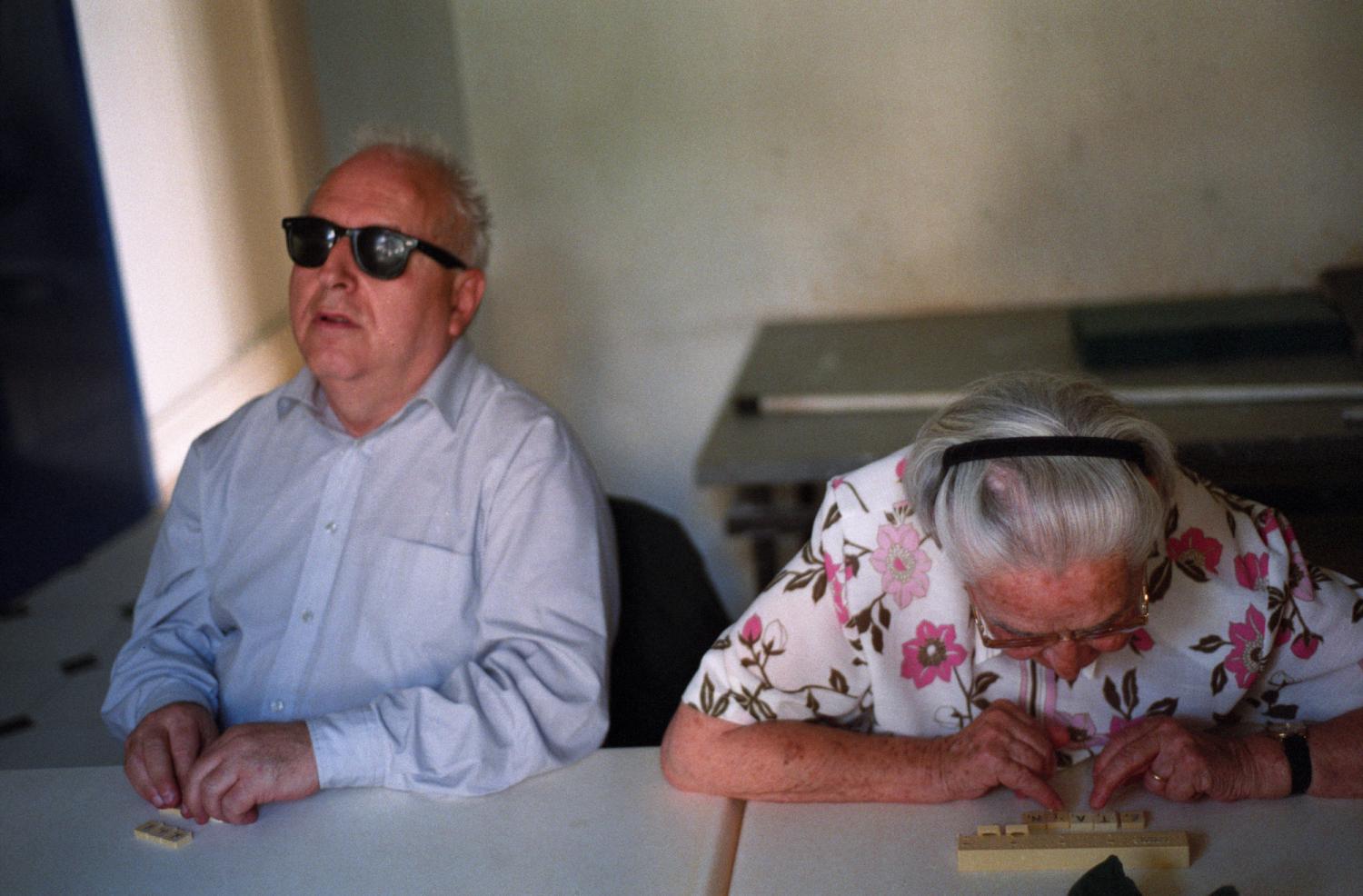 Bruno, lunettes noires et chemise bleues, lit les lettres de ses pions de scrabble avec les mains, tandis qu'à sa gauche Madeleine en chemise rose se penche vers les siens pour les déchiffrer avec les yeux. Paris, octobre 2005.