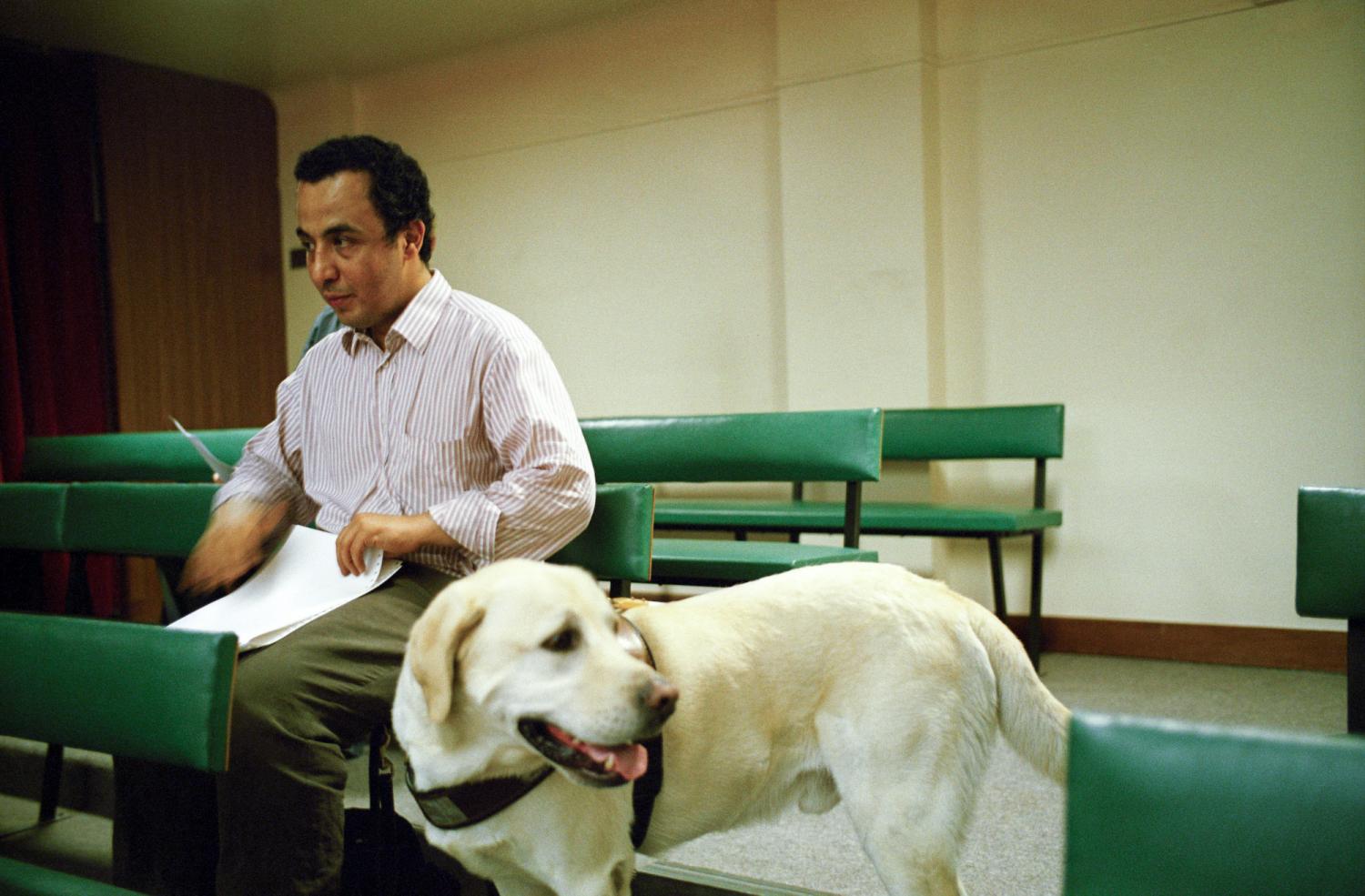 Assis sur un des petits bancs verts de la salle de spectacles de l'Association Valentin Haüy, près de son chien guide debout dans l'allée, Ali lit son programme en braille. Paris, juin 2005.