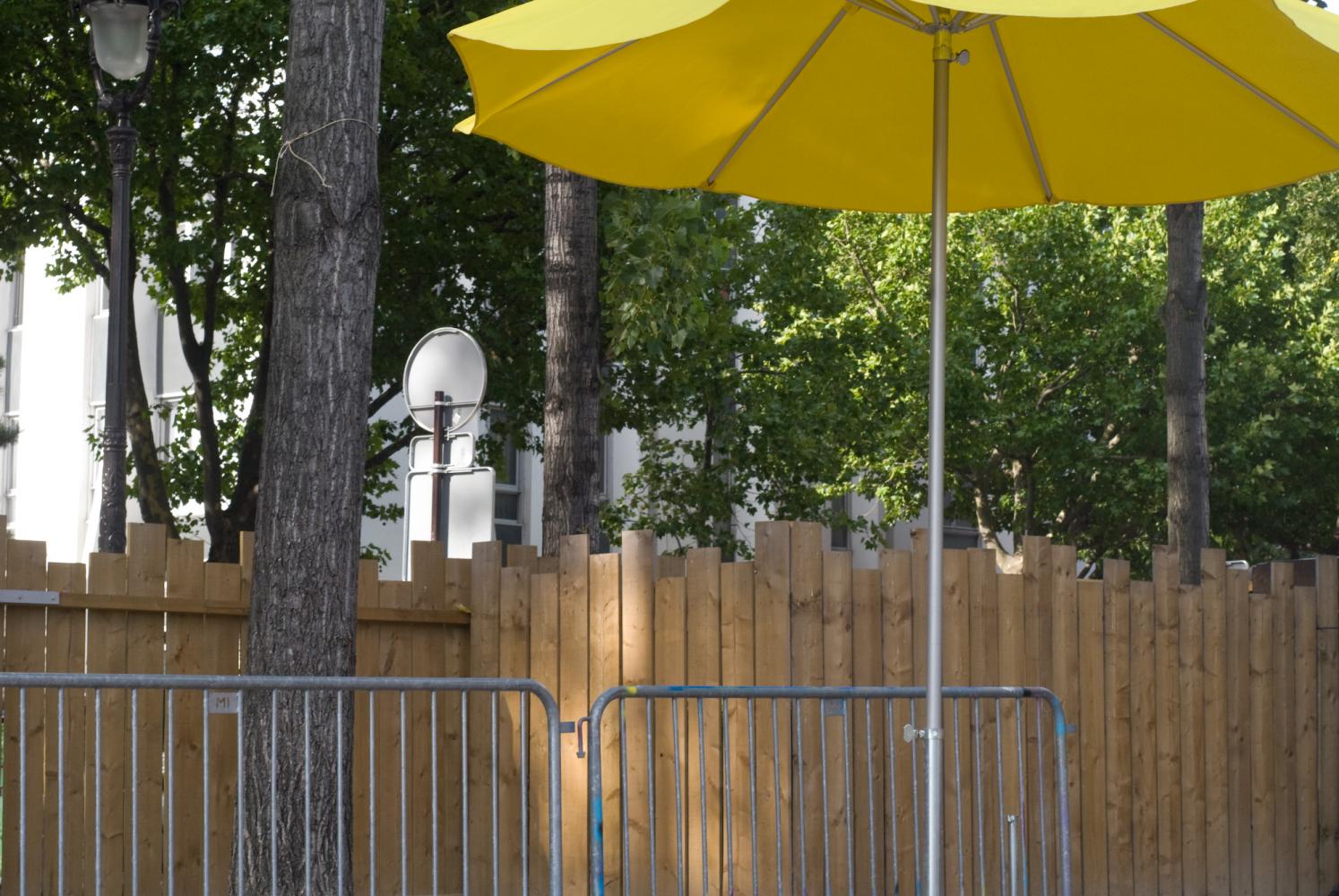 Un parasol jaune, des barrières métalliques, et derrière, une palissade se faufile entre les arbres. Paris, août 2010.