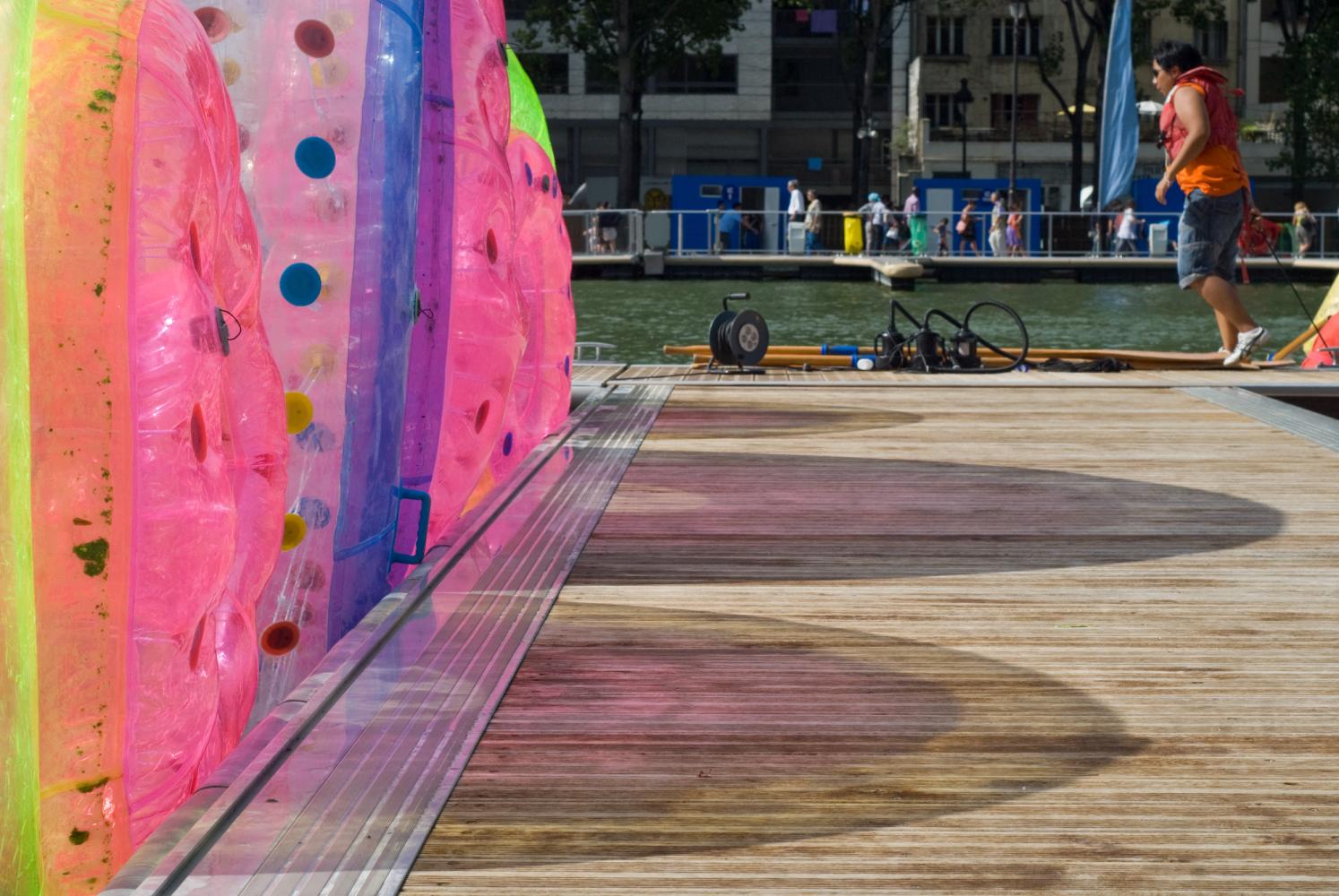 Collées au ponton, trois roues gonflables projettent leurs ombres colorées. Plus loin, une jeune personne en gilet de sauvetage s'apprête à descendre dans une petite embarcation. Paris, août 2010.