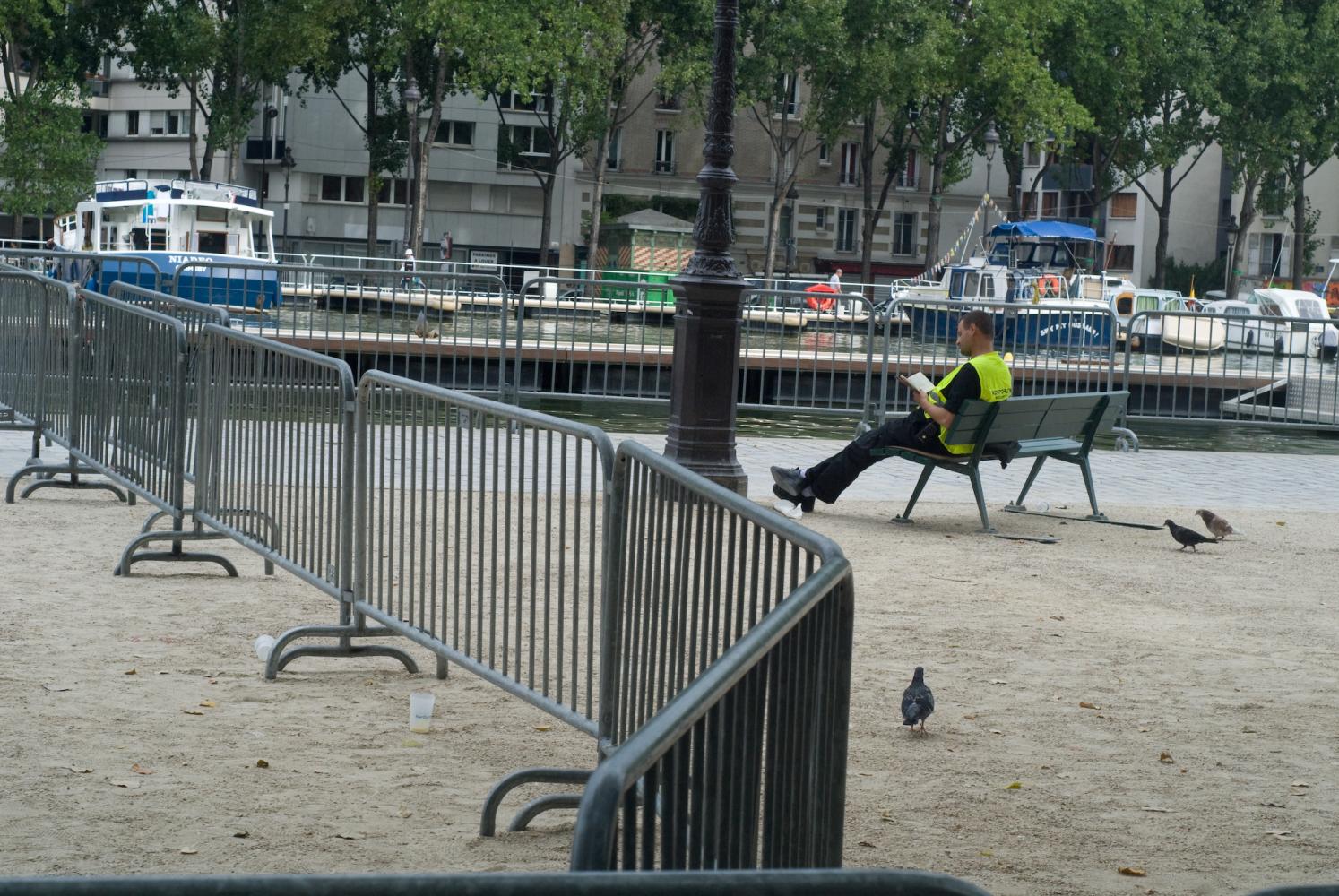 Assis sur un banc, cerné par des barrières métalliques, un homme en gilet jaune lit un livre parmi les pigeons. Paris, août 2010.