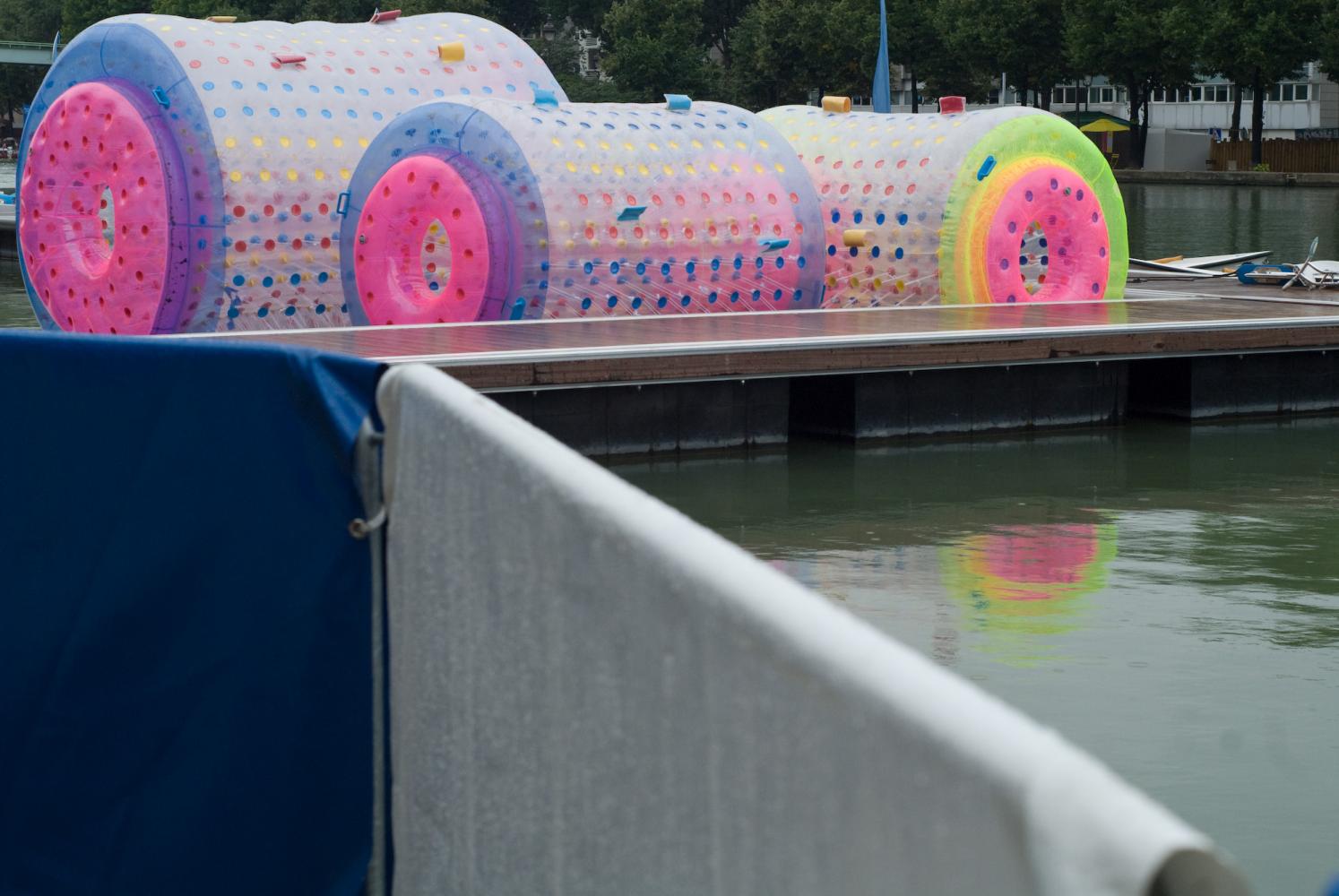 Amarrés à un ponton, derrière deux barrières métalliques recouvertes d'une bâche bleue et d'une blanche, trois roues gonflables aux couleurs vives. Paris, août 2010.