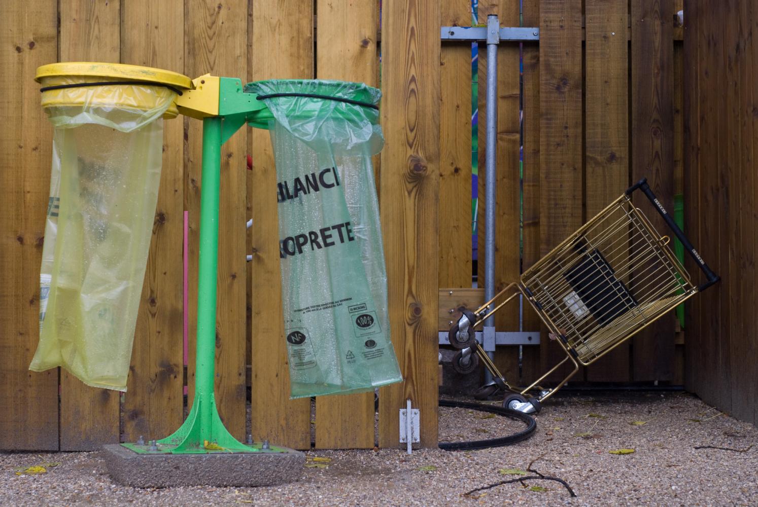 Devant une palissade en lattes de bois, une double poubelle, jaune et verte : deux sacs plastique et leur support, pour le recyclage. Derrière, dans un recoin, un caddie renversé. Paris, août 2010.
