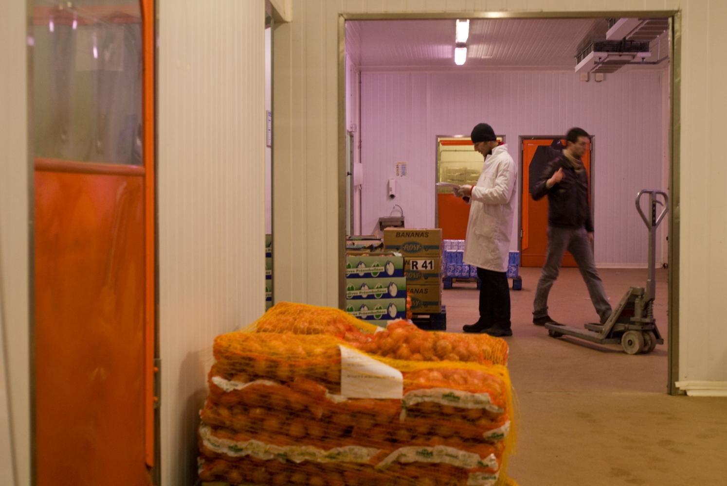Deux hommes dans un entrepôt, parmi des sacs d'oignons, des cartons d'endives ou de bananes et un transpalette. Rungis, mars 2009.