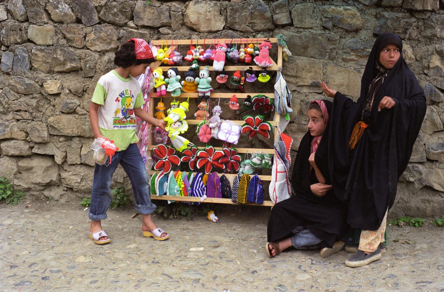 Une jeune visiteuse observe un stand de souvenirs tricotés tenu par deux fillettes en tchador. Masuleh, Iran, août 2006.