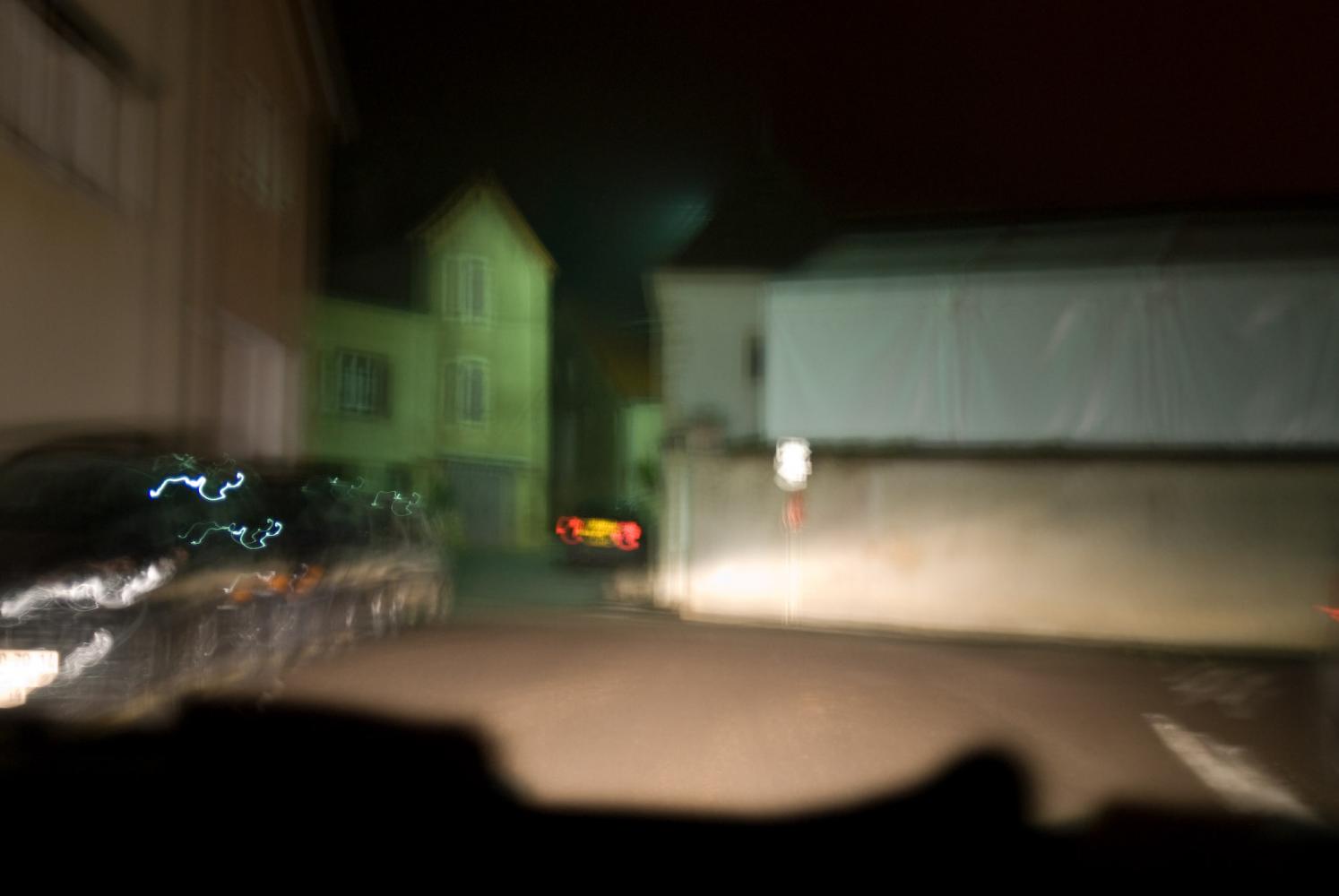 Une ruelle du village éclairée par des phares, les lumières rouges d'une autre voiture... tout un peu flou au retour de la paulée. Chassagne-Montrachet, Bourgogne, septembre 2009.