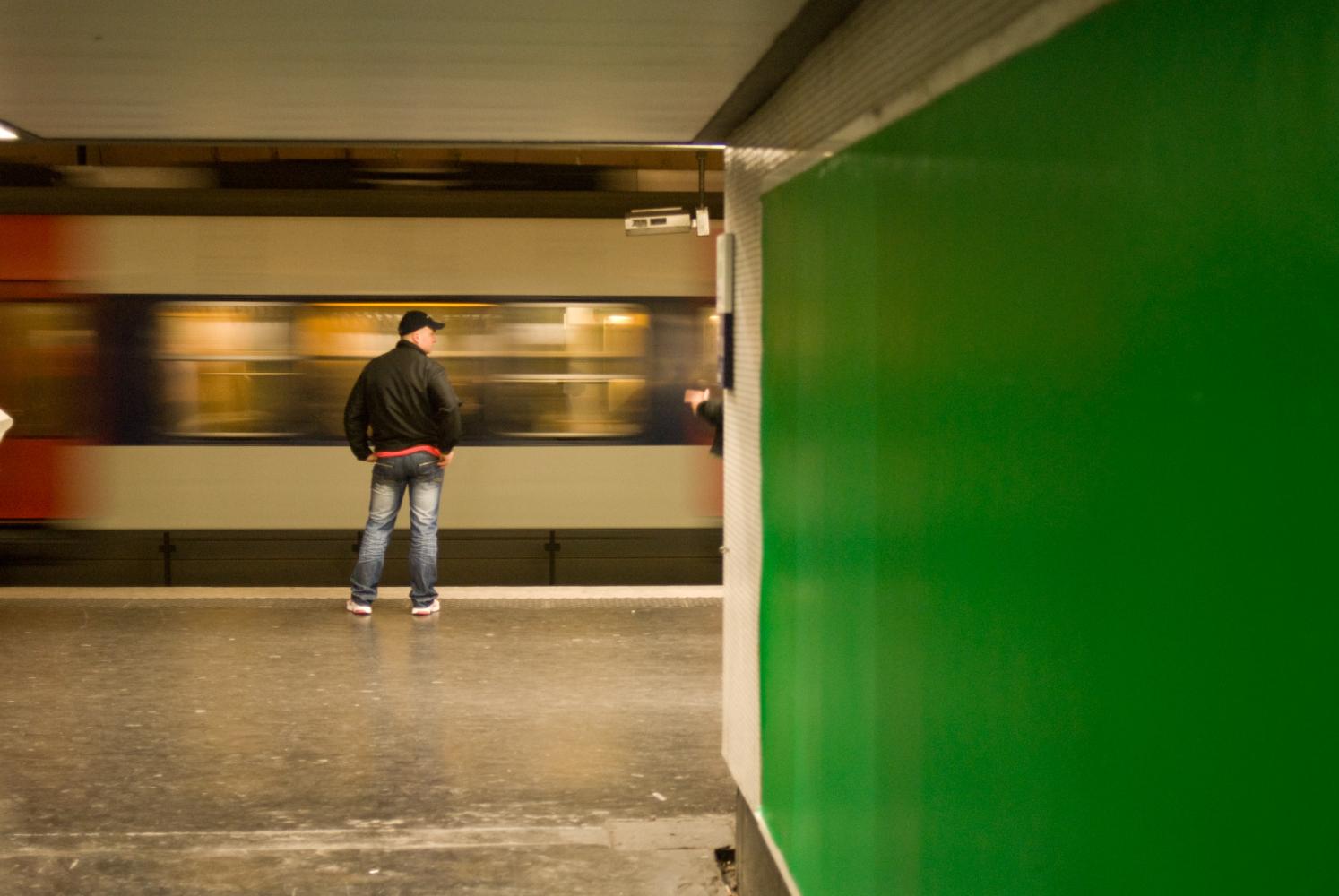 Sur le quai de RER à Nation, derrière le mur vert d'un couloir, un homme en jeans et casquette regarde passer un train. Paris, novembre 2009.