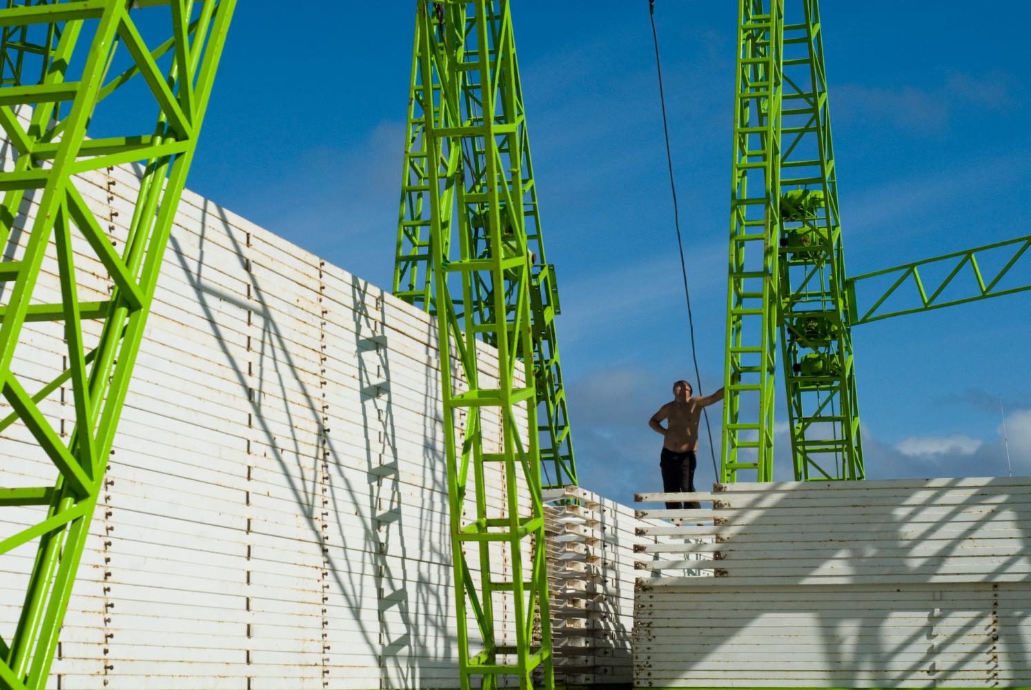 Debout sur des piles de barrières métalliques blanches, sur fond de ciel bleu, un homme s'appuie à la structure verte d'une grande roue foraine. Ténérife, Canaries, janvier 2010.